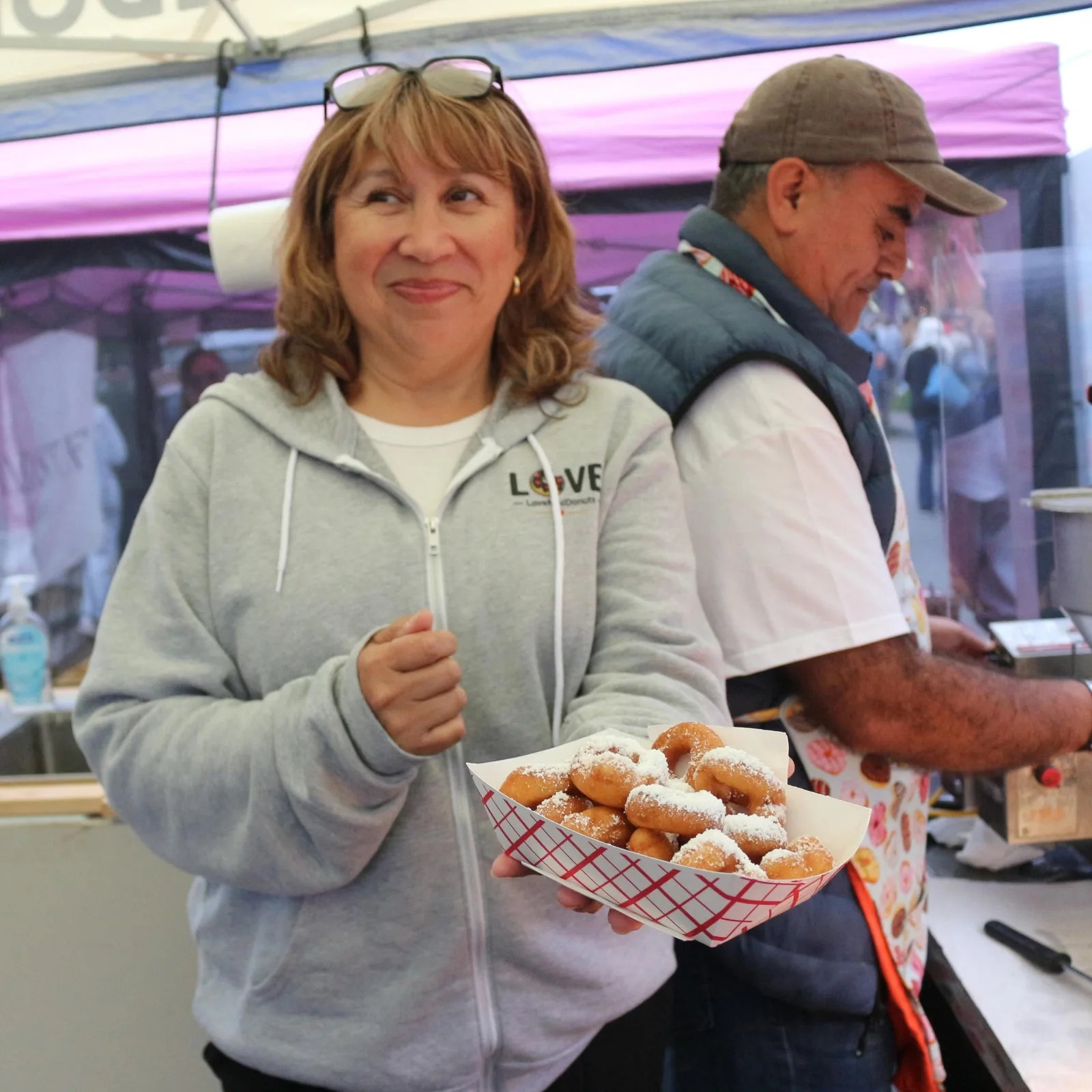Behind the counter under the tent: a tray of powdered mini donuts held out to the camera, the fryer working in the background.